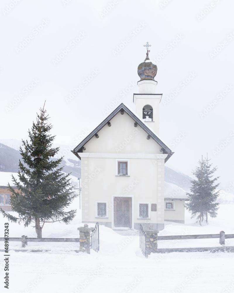 Small church of the Immaculate of Viera in Livigno, Italy in severe snow storm.  Snow blizzard and mountain in background, minimalist composition