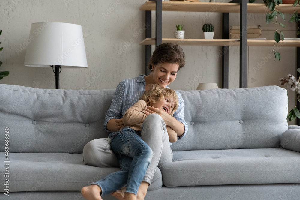 Affectionate happy millennial mother tickling laughing adorable small child daughter, relaxing together on comfortable sofa, enjoying carefree playtime weekend activity together on weekend at home. Affectionate happy millennial mother tickling laughing adorable small child daughter, relaxing together on comfortable sofa, enjoying carefree playtime weekend activity together on weekend at home.