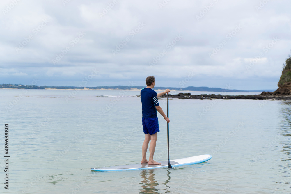 Man wearing rash guard on a stand-up paddle board at the ocean bay in Australia. SUP water sport activity Man wearing rash guard on a stand-up paddle board at the ocean bay in Australia. SUP water sport activity