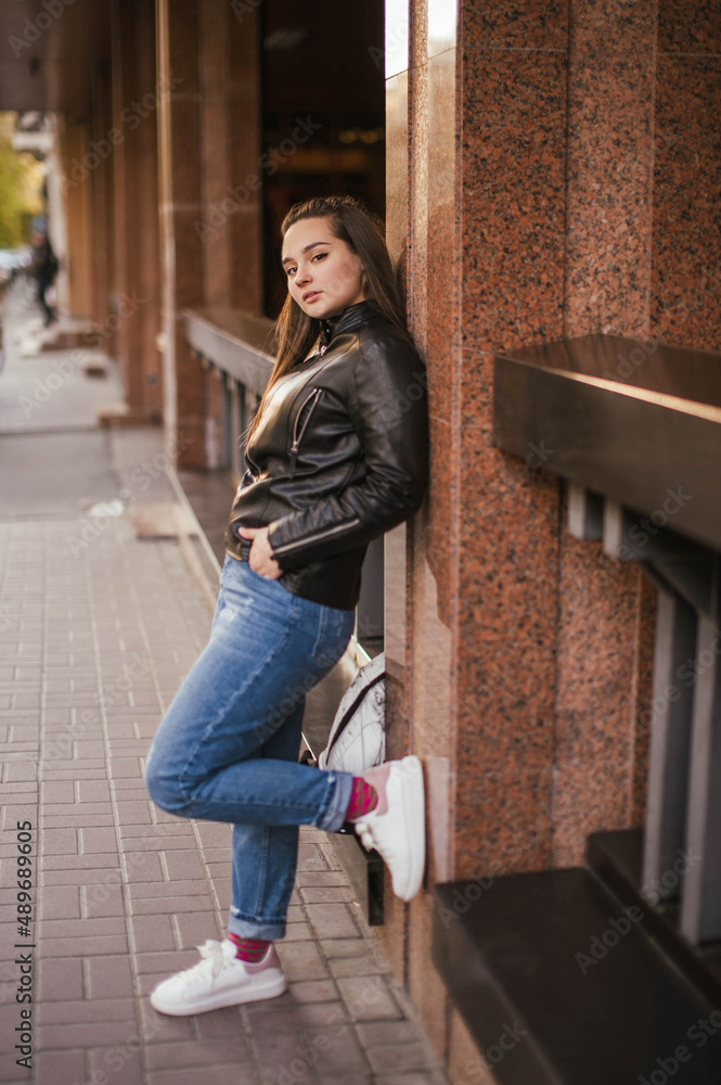 Photo of a brown-haired girl in a leather jacket and jeans on the street in the city