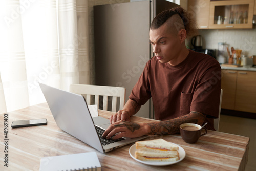 Serious young man having coffee and sandwich for breakfast when working on laptop at table in kitchen