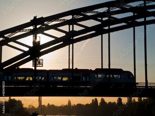 Silhouette eines über eine Metallbogenbrücke fahrenden Personenzugs im Gegenlicht der aufgehenden Sonne