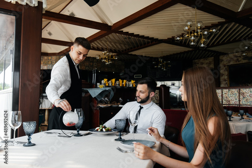 A young waiter in a stylish apron serves a table with a beautiful couple in an elegant restaurant. Customer service in an elite restaurant and public catering establishment.
