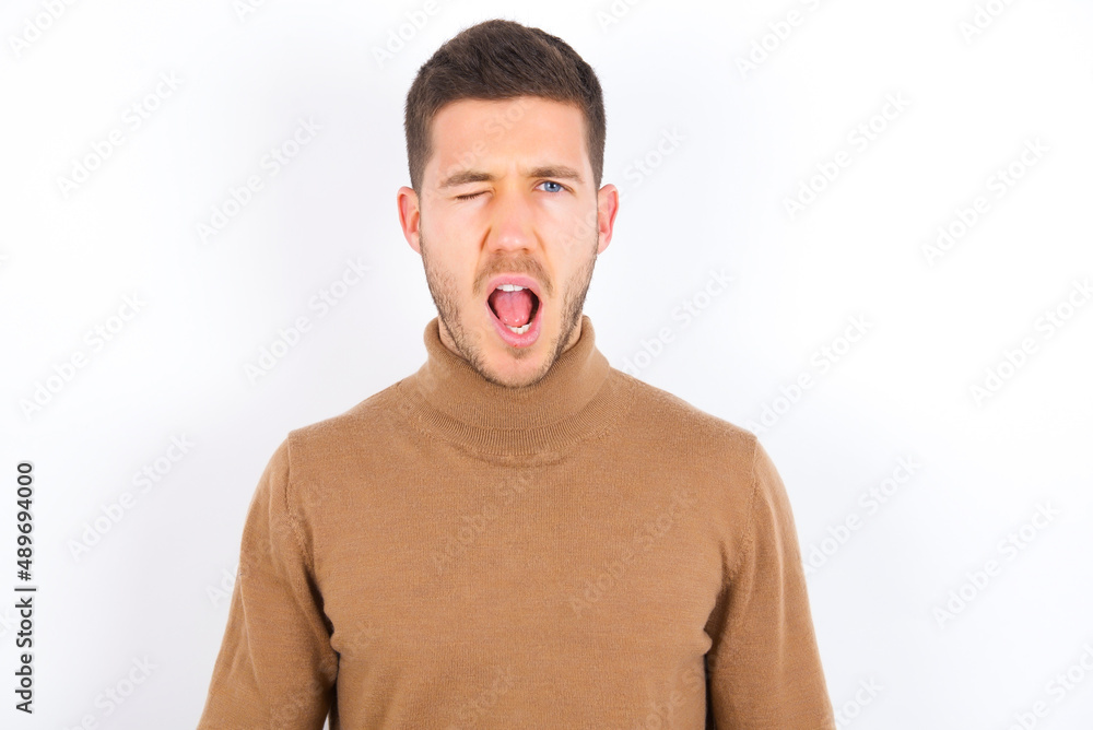 young caucasian man wearing grey turtleneck over white background winking looking at the camera with sexy expression, cheerful and happy face. young caucasian man wearing grey turtleneck over white background winking looking at the camera with sexy expression, cheerful and happy face.