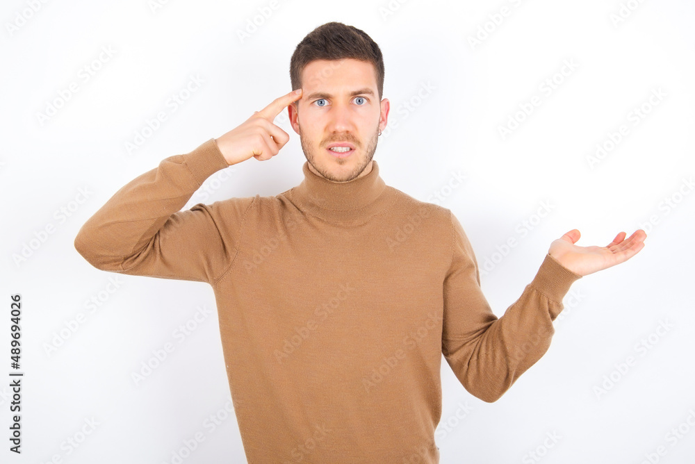 young caucasian man wearing grey turtleneck over white background confused and annoyed with open palm showing copy space and pointing finger to forehead. Think about it. young caucasian man wearing grey turtleneck over white background confused and annoyed with open palm showing copy space and pointing finger to forehead. Think about it.
