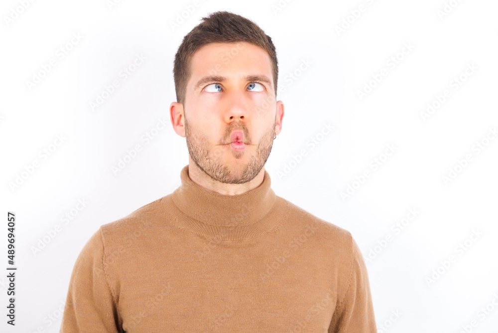 Fototapeta premium young caucasian man wearing grey turtleneck over white background making fish face with lips, crazy and comical gesture. Funny expression.