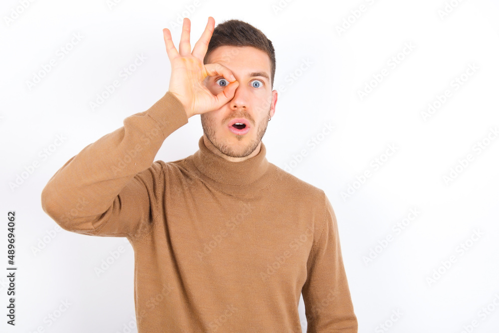 young caucasian man wearing grey turtleneck over white background doing ok gesture shocked with surprised face, eye looking through fingers. Unbelieving expression. young caucasian man wearing grey turtleneck over white background doing ok gesture shocked with surprised face, eye looking through fingers. Unbelieving expression.