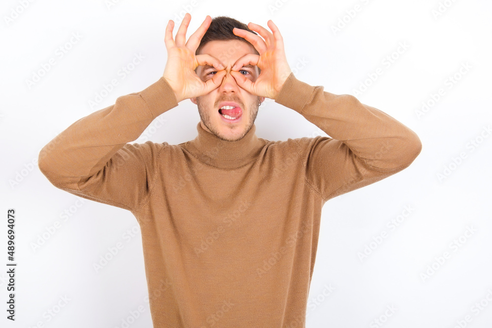 young caucasian man wearing grey turtleneck over white background doing ok gesture like binoculars sticking tongue out, eyes looking through fingers. Crazy expression. young caucasian man wearing grey turtleneck over white background doing ok gesture like binoculars sticking tongue out, eyes looking through fingers. Crazy expression.