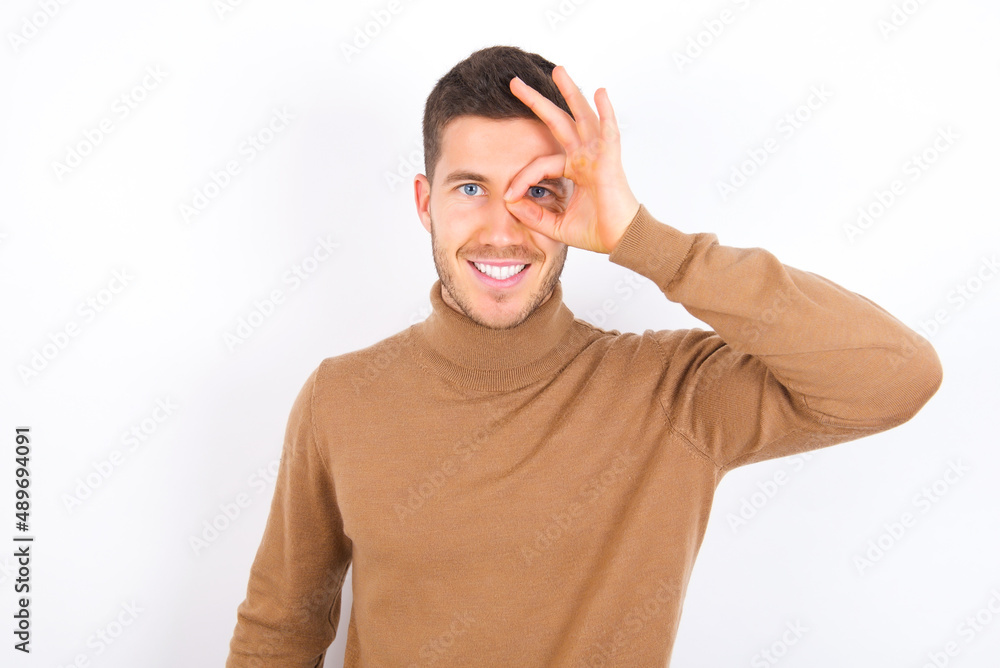 young caucasian man wearing grey turtleneck over white background doing ok gesture with hand smiling, eye looking through fingers with happy face. young caucasian man wearing grey turtleneck over white background doing ok gesture with hand smiling, eye looking through fingers with happy face.