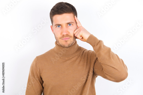 young caucasian man wearing grey turtleneck over white background tries to memorize something, keeps fore finger on temple, reminds information for exam,