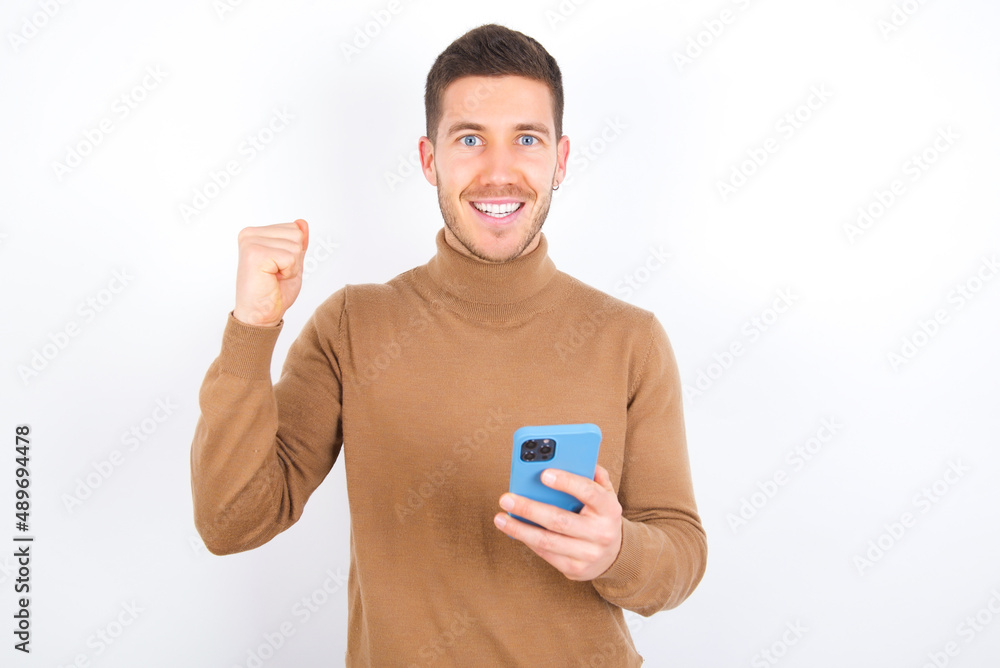Portrait of young caucasian man wearing grey turtleneck over white background holding in hands cell celebrating Portrait of young caucasian man wearing grey turtleneck over white background holding in hands cell celebrating