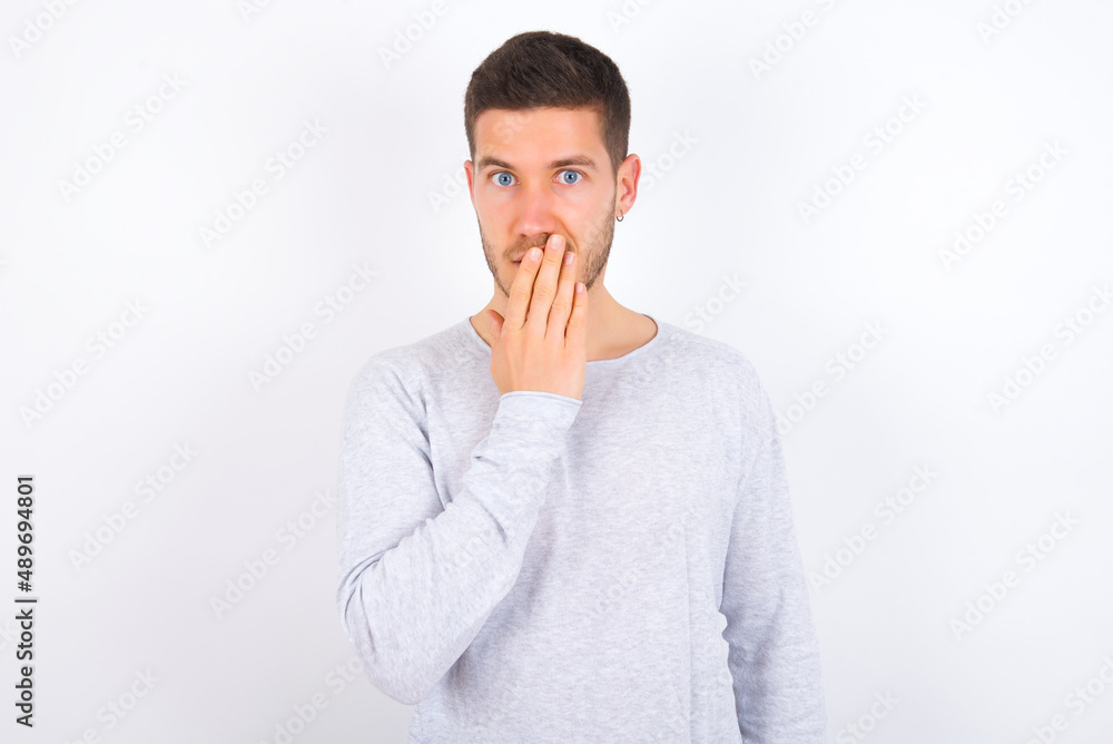 young caucasian man wearing grey sweater over white background covers mouth and looks with wonder at camera, cannot believe unexpected rumors. young caucasian man wearing grey sweater over white background covers mouth and looks with wonder at camera, cannot believe unexpected rumors.