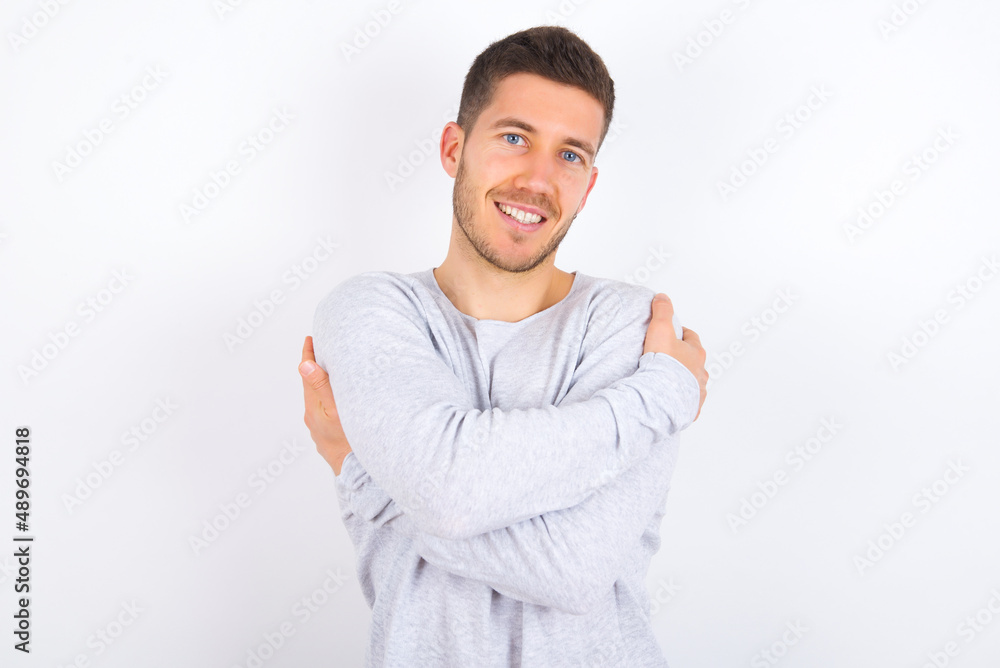 Charming pleased young caucasian man wearing grey sweater over white background embraces own body, pleasantly feels comfortable poses. Tenderness and self esteem concept Charming pleased young caucasian man wearing grey sweater over white background embraces own body, pleasantly feels comfortable poses. Tenderness and self esteem concept