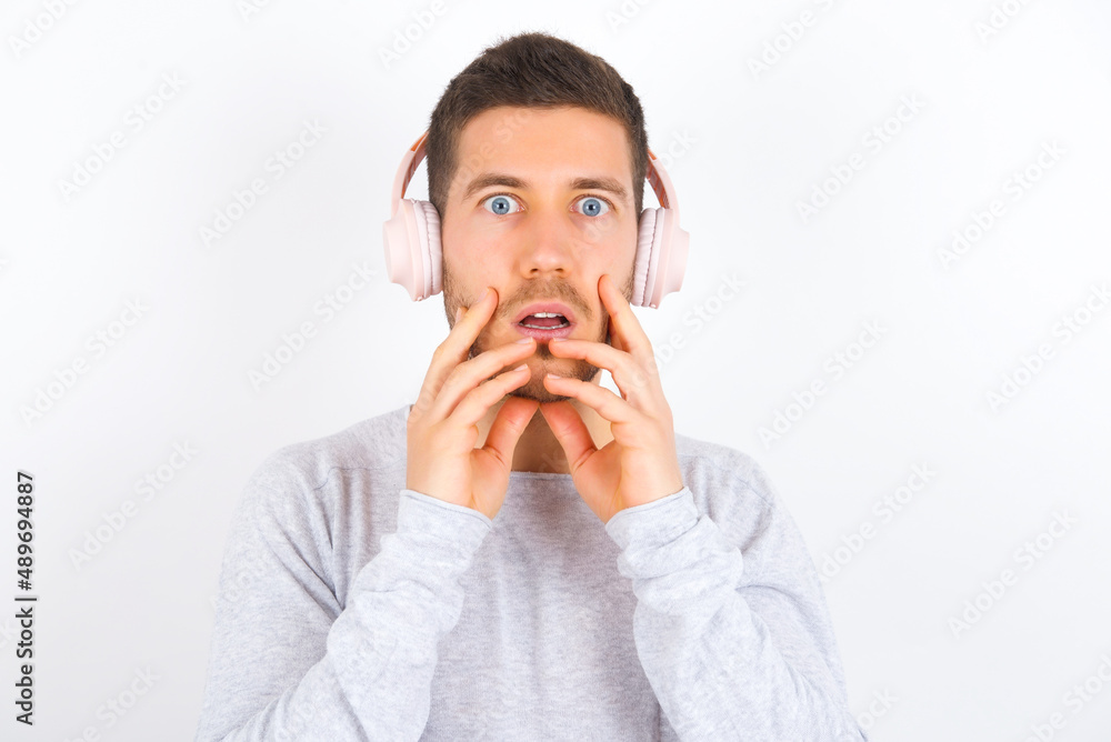 Shocked young caucasian man wearing grey sweater over white background stares fearful at camera keeps mouth widely opened wears wireless stereo headphones on ears Shocked young caucasian man wearing grey sweater over white background stares fearful at camera keeps mouth widely opened wears wireless stereo headphones on ears