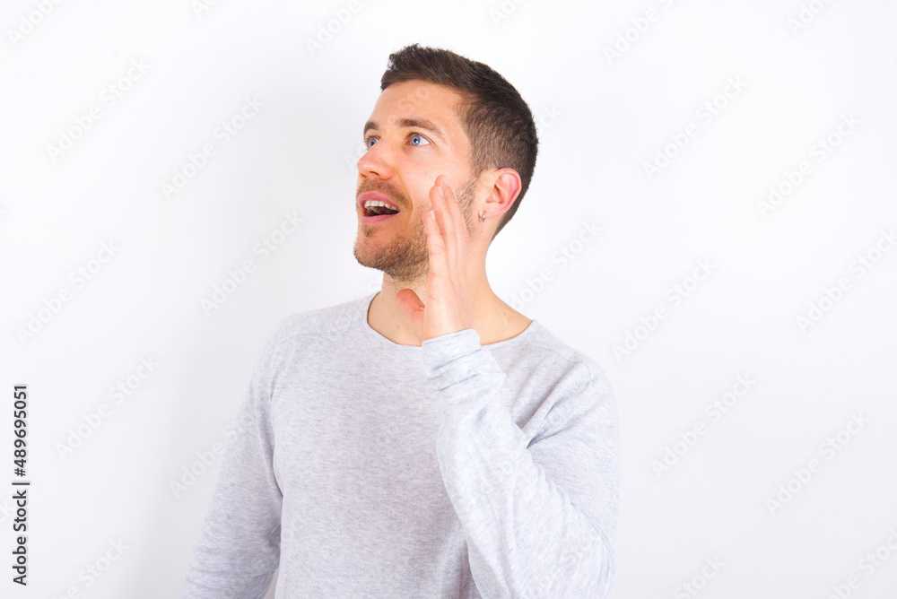young caucasian man wearing grey sweater over white background look empty space holding hand near her face and screaming or calling someone. young caucasian man wearing grey sweater over white background look empty space holding hand near her face and screaming or calling someone.