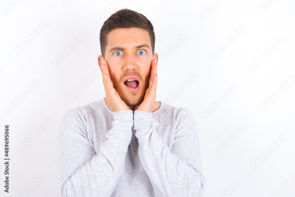 Upset young caucasian man wearing grey sweater over white background touching face with two hands Upset young caucasian man wearing grey sweater over white background touching face with two hands