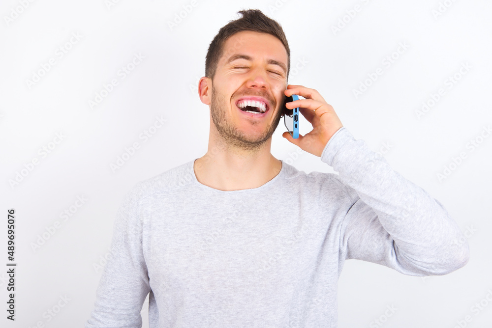 Overemotive happy young caucasian man wearing grey sweater over white background laughs out positively hears funny story from friend during telephone conversation Overemotive happy young caucasian man wearing grey sweater over white background laughs out positively hears funny story from friend during telephone conversation