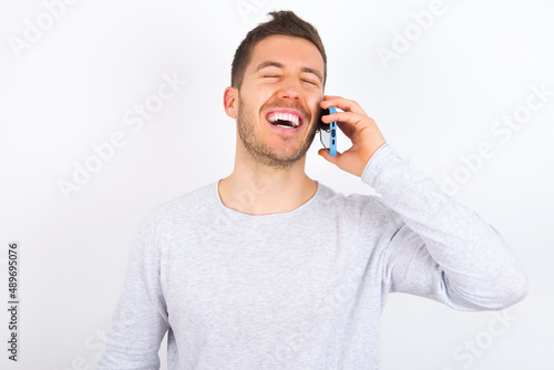 Overemotive happy young caucasian man wearing grey sweater over white background laughs out positively hears funny story from friend during telephone conversation