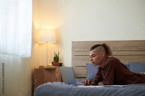 Young man lying on bed, working on laptop, coding or andswering e-mails