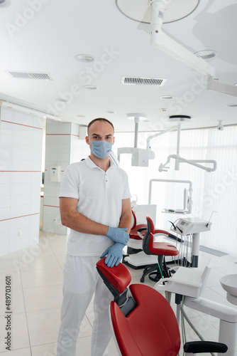 A young dentist in a mask stands near a red dental chair and smiles in modern white dentistry. Modern dentistry and prosthetics.