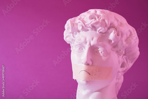 copy of the head of an antique statue of David with a taped mouth in pink neon light on a purple background © Natasha