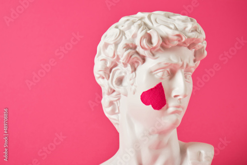a copy of the head of an antique statue of David with a pasted pink heart on his cheek, a bright pink background