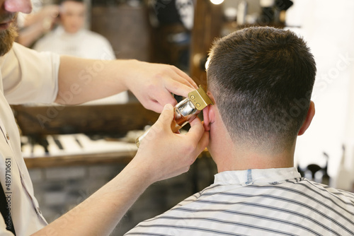 Professional male hairdresser does hairstyle to bearded man in barber shop, back view, barber's hand with clipper.
