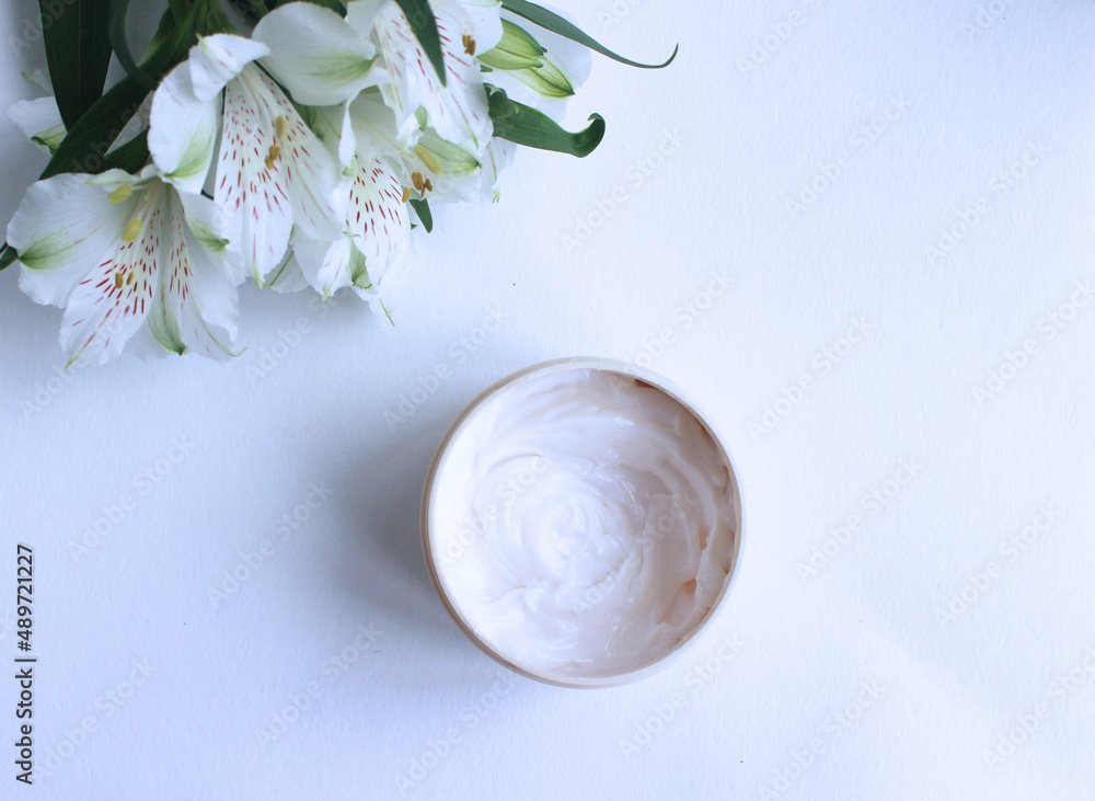 White flowers of alstroemeria and a greasy cosmetic cream in a round jar on a white background. Top view. White flowers of alstroemeria and a greasy cosmetic cream in a round jar on a white background. Top view.