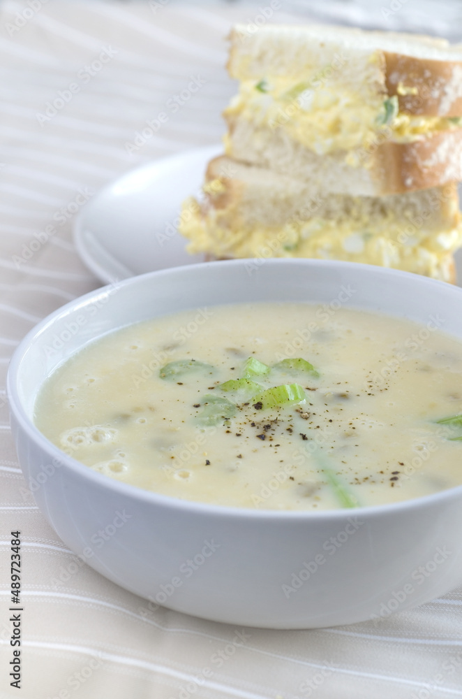 Cream of Celery Soup in a White Bowl with Blurred Egg Salad Sandwich in Background