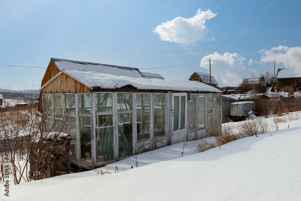 Homemade greenhouse from glass and wood in snow in russian dacha in sunny winter day. The greenhouse in the garden is covered with snow.