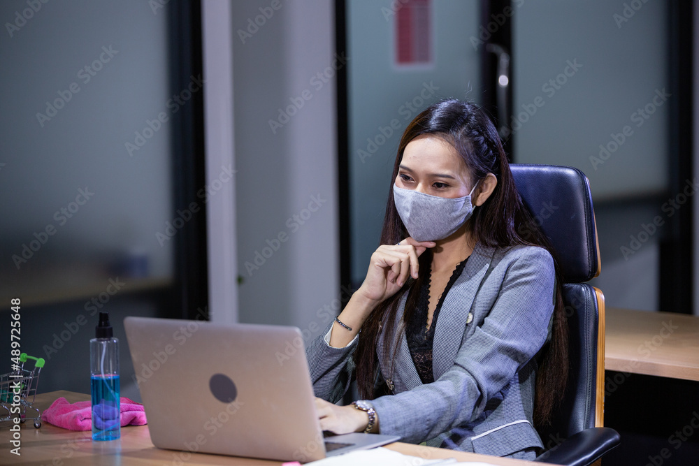Asian woman in protective mask using laptop working overtime at desk in the office. serious thinking of work. browsing and searching data for project Asian woman in protective mask using laptop working overtime at desk in the office. serious thinking of work. browsing and searching data for project