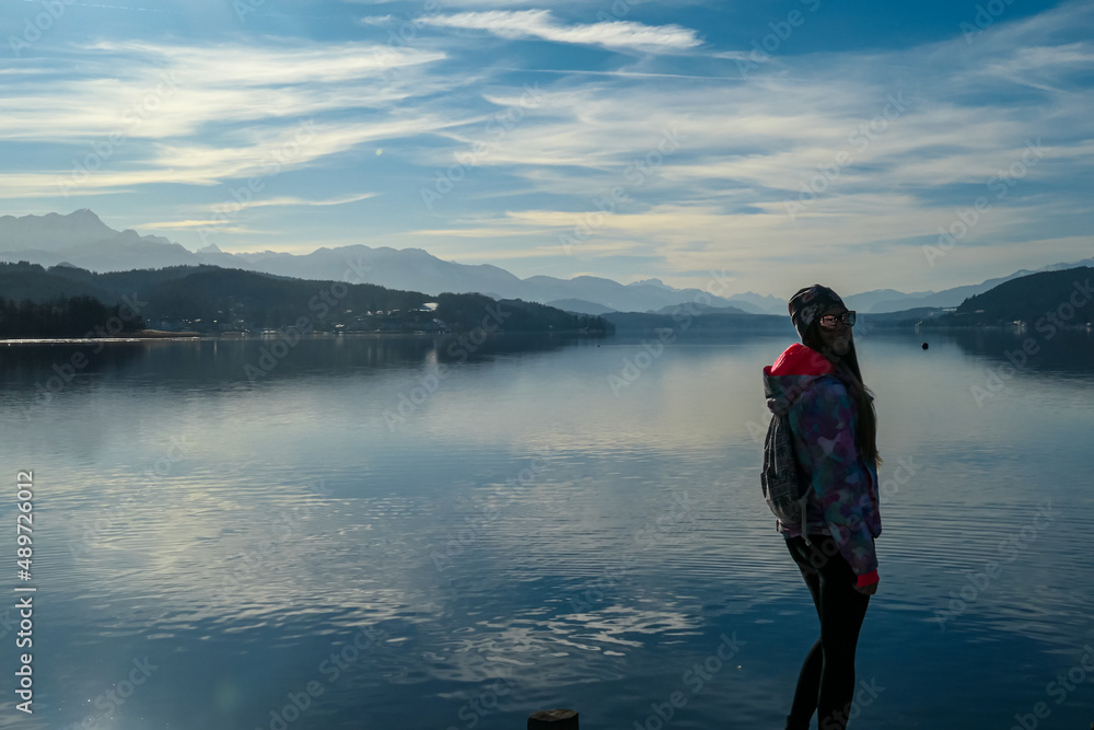 Happy woman standing on the pier at beautiful Woerthersee in Poertschach, Carinthia, Austria. Scenic lake landscape surrounded by Karawanks Alps. Fresh and clean air. Reflection in Lake Woerth