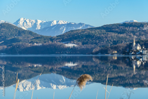 View on Maria Worth and Woerthersee from Poertschach in Carinthia, Austria. Calm lake reflecting the landscape. View on Koschutnikturm and Hochobir. Village with church at the waters edge. Reed photo