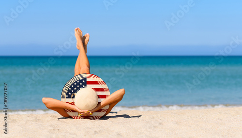 Fototapeta Naklejka Na Ścianę i Meble -  A slender girl on the beach in a straw hat in the colors of the USA flag. The concept of a perfect vacation in a resort in the United States. Focus on the hat.