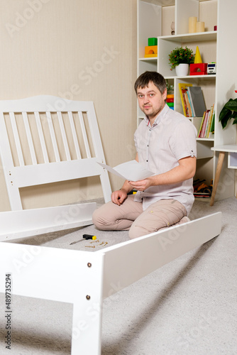 A man with instructions for assembling furniture is going to assemble a children's white wooden crib in a children's bedroom.