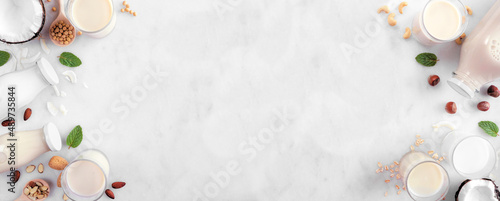 Vegan, plant based, non dairy milk double border. Assortment of types in milk bottles and glasses with scattered ingredients. Overhead view on a white marble background with copy space.