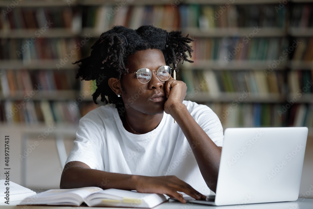 Unhappy young African American hipster guy in eyeglasses looking in distance, feeling bored preparing for examination or doing high school research project on computer, sitting at table in library. Unhappy young African American hipster guy in eyeglasses looking in distance, feeling bored preparing for examination or doing high school research project on computer, sitting at table in library.