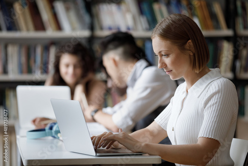 Happy focused smart Caucasian female student using computer writing essay or coursework, working on high school project, preparing for exams, sitting at shared table in public or college library.