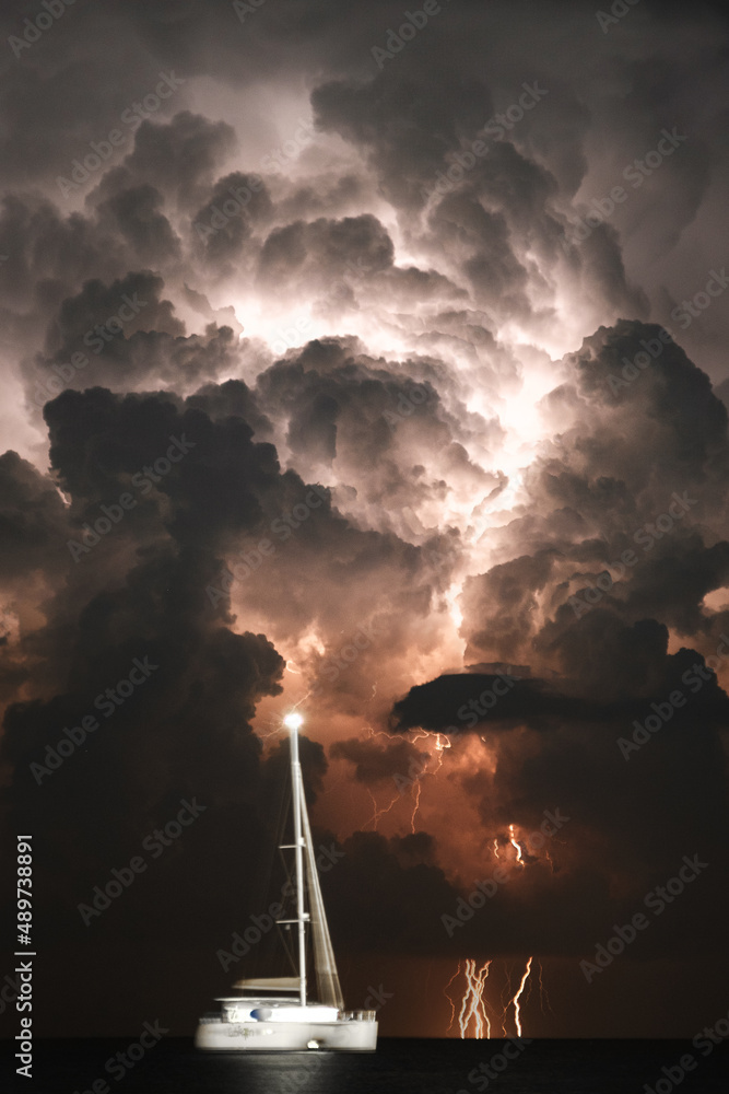 nächtliche leuchtende Gewitterfront mit Blitzen und ankerndem Segelboot im Vordergrund