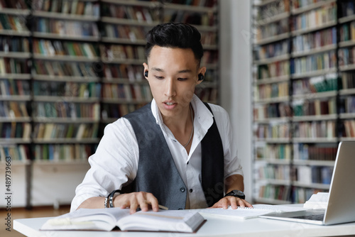 Concentrated millennial male Korean student in earbuds listening favorite lounge music or educational lecture while reading book, preparing for exam or doing research alone in college library.