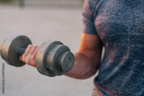 Close-up arm of unrecognizable athletic male with strong muscular body lifting dumbbell during summer evening working out outdoor. Sportsman pumping muscles with weight in city park, selective focus.