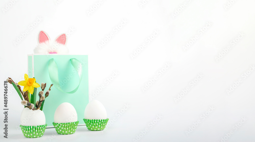 Three white unpainted Easter eggs in green paper cake pans with yellow narcissus and willow branches and green gift bag with funny bunny ears on white background. Easter spring concept. Copy space