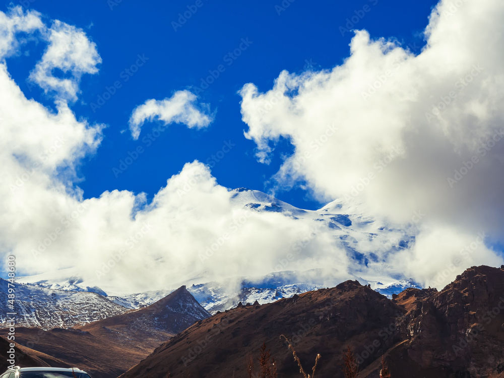 In a rocky autumn mountain valley, the white snowy peak of Elbrus is visible behind the clouds. Caucasus mountains under a bright blue sky