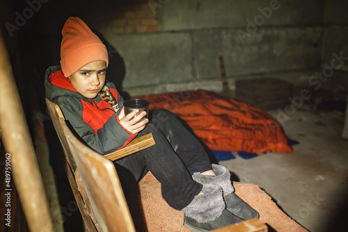 Terrorism and war, current history. Little Ukrainian girl sits in a bomb shelter, drinks tea from a thermos and waits for the end of the air attack of Russian invaders photo