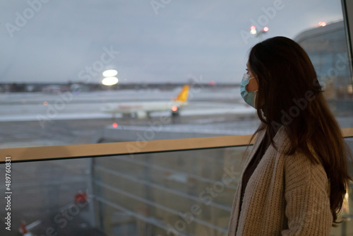 A Caucasian woman is standing near the window in airport and looking at plane