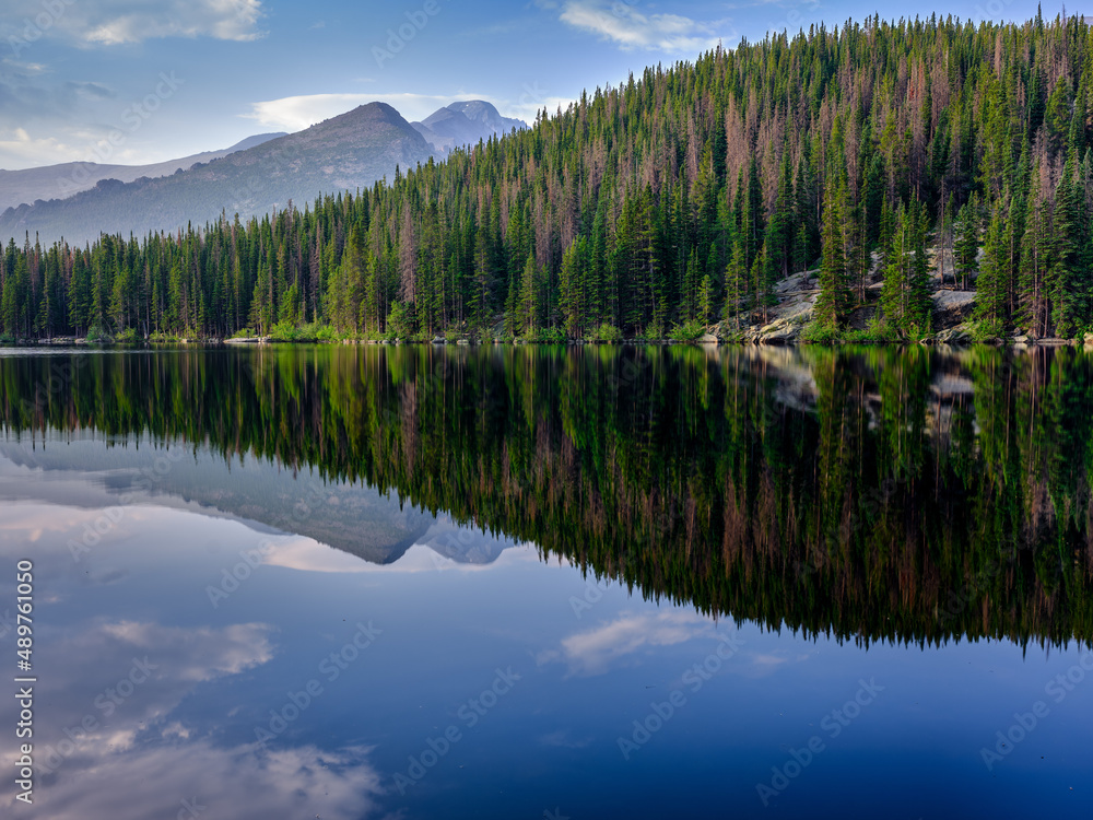 Pine trees reflected in the clear and calm waters of Bear Lake in the Colorado Rocky Mountains with mountain peaks in the background
