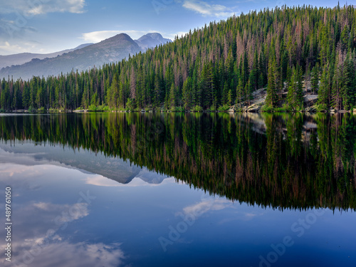 Pine trees reflected in the clear and calm waters of Bear Lake in the Colorado Rocky Mountains with mountain peaks in the background