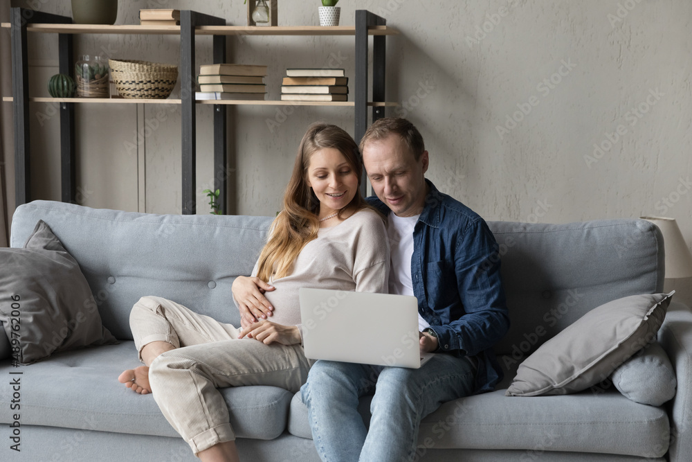 Happy family pregnant couple relaxing on comfortable couch together, using computer, shopping with online ecommerce app on laptop, browsing Internet, talking on video call, touching big belly Happy family pregnant couple relaxing on comfortable couch together, using computer, shopping with online ecommerce app on laptop, browsing Internet, talking on video call, touching big belly