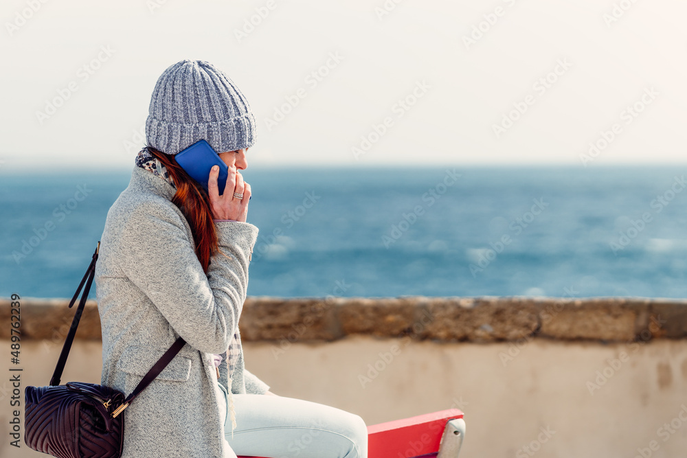 red-haired woman in woollen cap talking on mobile phone looking out to the ocean