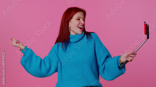 Happy teenager student adult girl blogger taking selfie on mobile phone selfie stick, dancing, communicating video call online with subscribers. Young fashionable woman on pink studio background